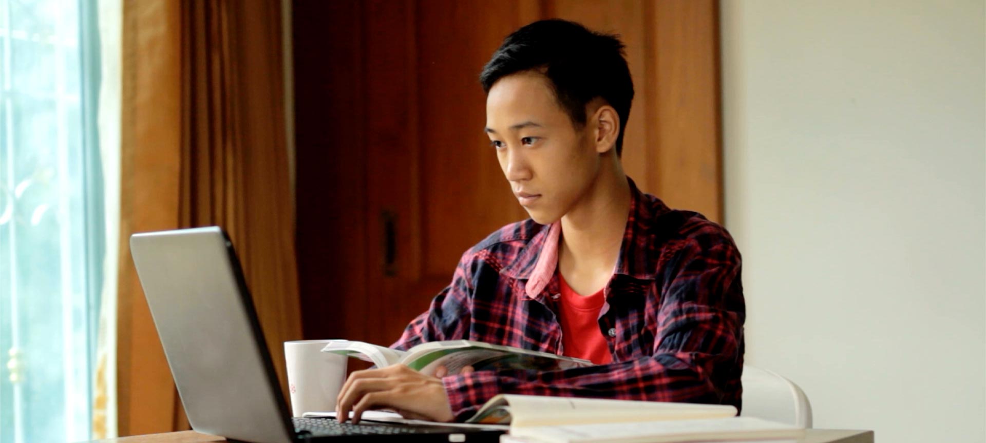 Male student using a laptop to study with book in hand
